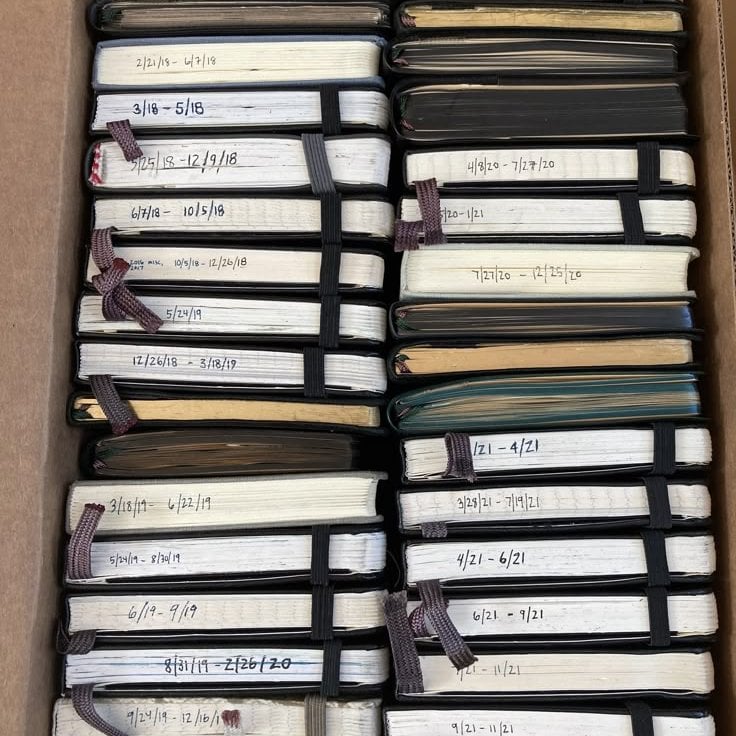 A box full of finished, dated, handwritten notebooks stacked in chronological order